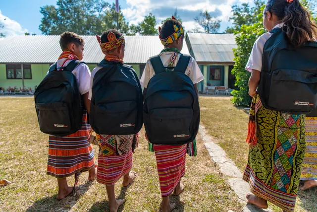 Four pupils in woven sarongs and traditional headbands walk towards school with new backpacks